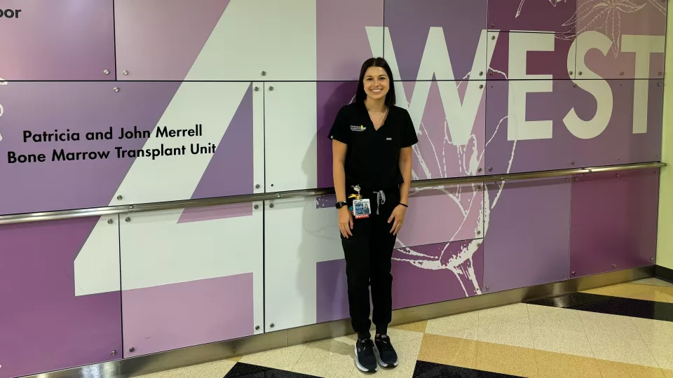 Kate, wearing black scrubs, stands smiling in front of the purple 4 West lobby wall