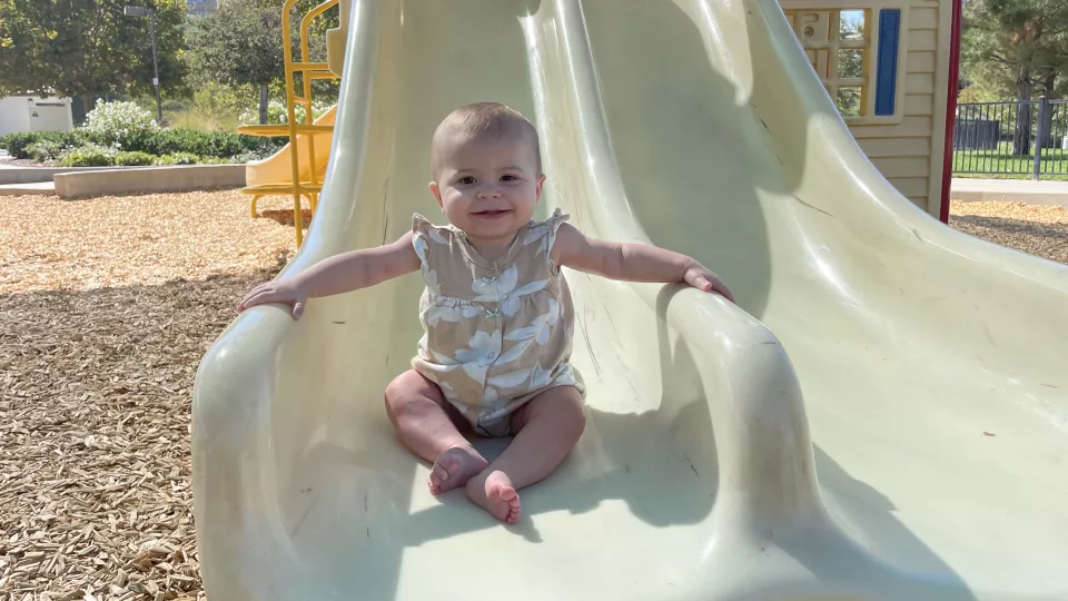 A light skin tone baby wearing a tan and white outfit, smiling and sitting on a slide at the playground