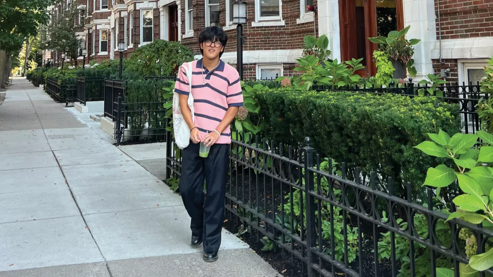 Teenage boy with medium skin tone and dark hair wearing a black and pink short sleeved collared shirt and long black pants, standing outside near a brick building.