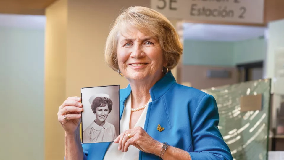 A woman with light skin tone, short blonde hair wearing a light blue jacket holding a black and white picture of her younger self.